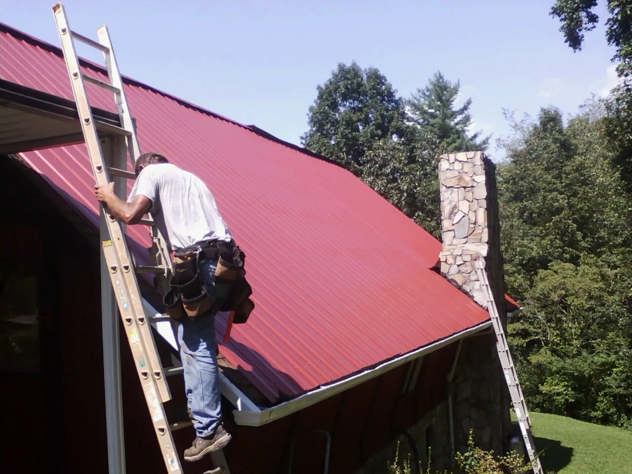 Roofer on a ladder beside a red metal roof and stone chimney — Soffit & Fascia Repair by Roofing Brothers in Hellier